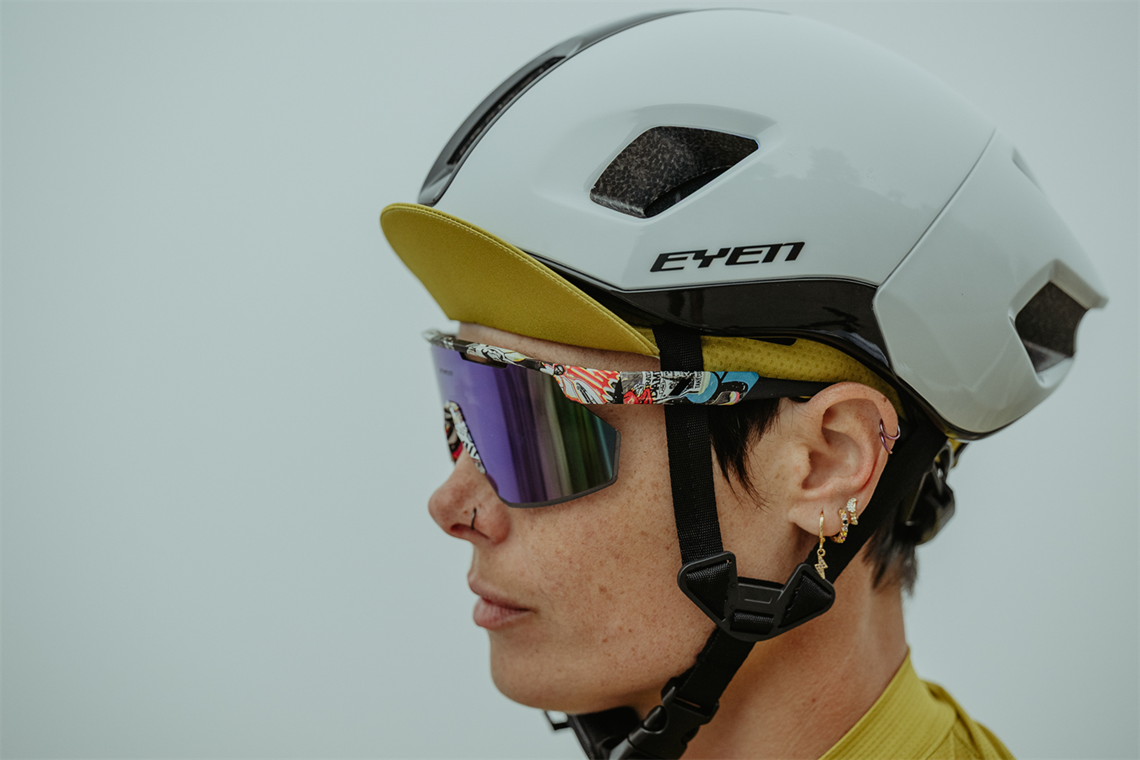 Kolarska stoi bokiem we mgle, ma ubrany aerodynamiczny kask, okulary z komiksową ramką, czapeczkę z daszkiem pod kaskiem i kilka kolczyków na uszach plus jeden na nosie.